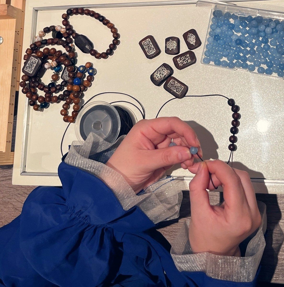 Artisane en caftan bleu enfilant un bracelet en perles d'oud, atelier Soltan Al Oud à Kénitra